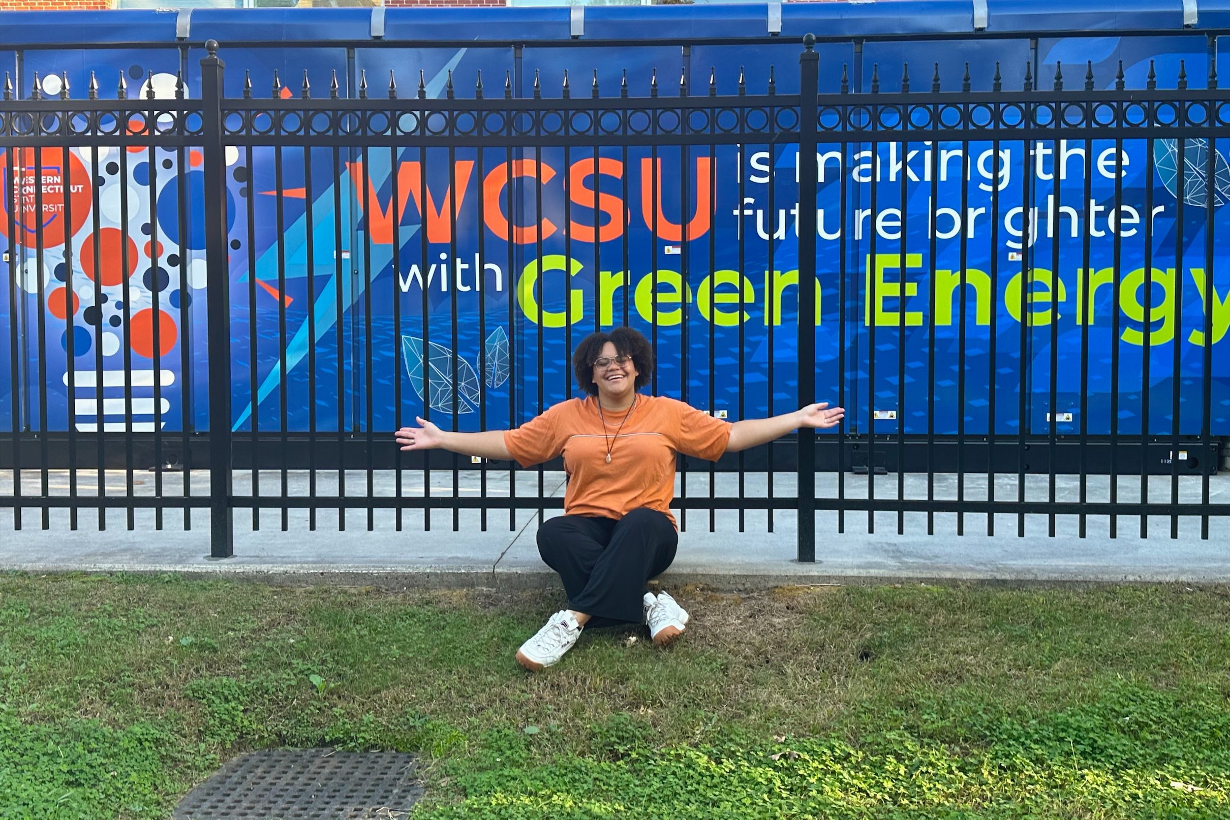 Denise Sweeney in an orange shirt and black pants with arms stretched out sitting infront of a black metal fence in front of a fuel cell generator that reads "WCSU is making the future brighter with green energy" on a blue background with a lightblub graphic and the WCSU logo